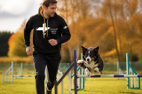 Handler in a Cornwall Agility Club hoodie running beside his dog over an agility jump