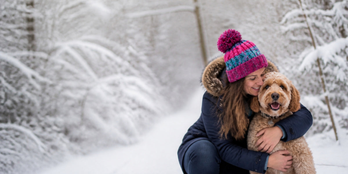 Woman in a pink and teal pom pom beanie hugging her Cockapoo on a snowy woodland walk
