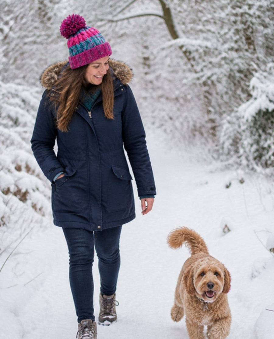 Woman in a pom pom beanie walking a Cockapoo through snowy woods