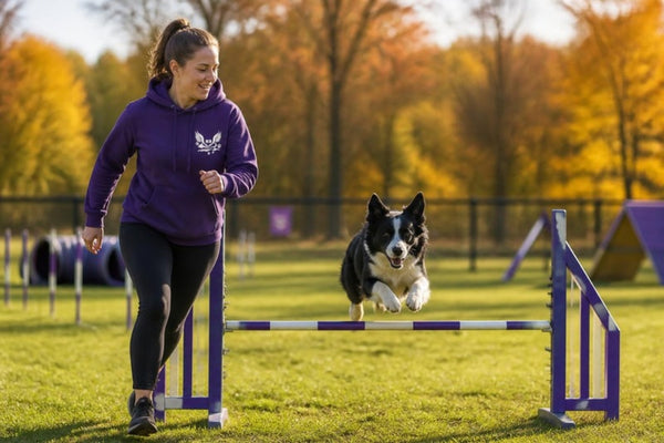 Handler in a purple K9 Heaven hoodie running beside her dog over an agility jump