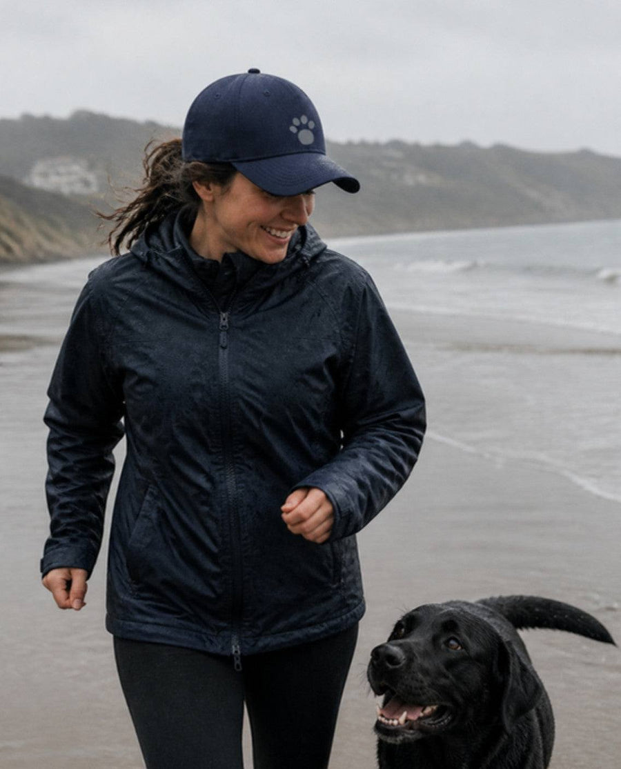 Woman in a navy paw print cap on a wet beach with a black Labrador